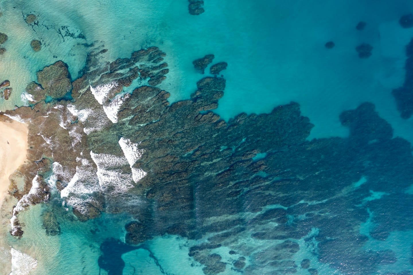 Turquoise ocean waves crash on dark rocks by a sandy beach.