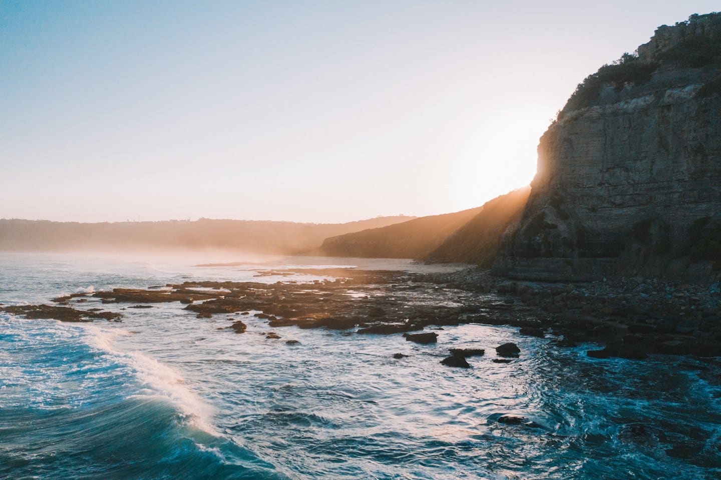 Waves crash against a dark cliff along a rugged coastline.